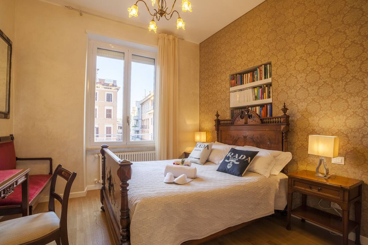 A hotel room with a bed, a bookshelf, a wooden nightstand, and a view of the city through the window.
