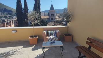 Varanda com mesa, duas cadeiras e um banco. Há plantas em vasos e vista para um edifício com torre.