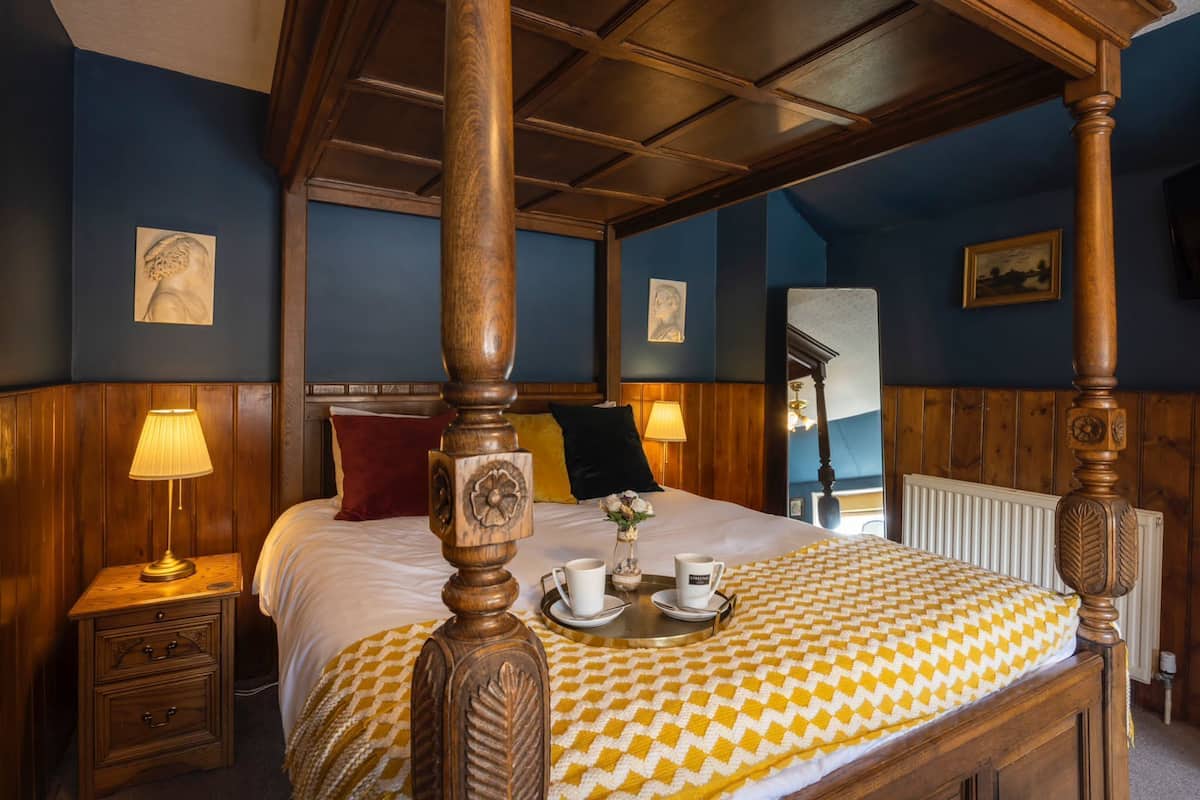 A four-poster bed with a checkered bedspread, bedside lamps, and a tray with cups and a flower.