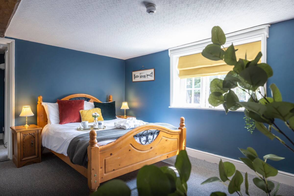 A bedroom with a wooden bed, two bedside tables with lamps, a window with a yellow blind, and a plant in the foreground.