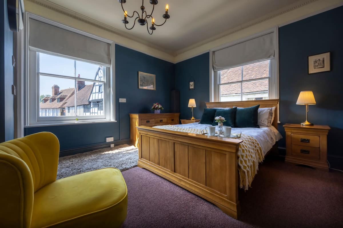 A bedroom with a large bed, a wooden nightstand, a yellow armchair, and a window with a view of buildings.