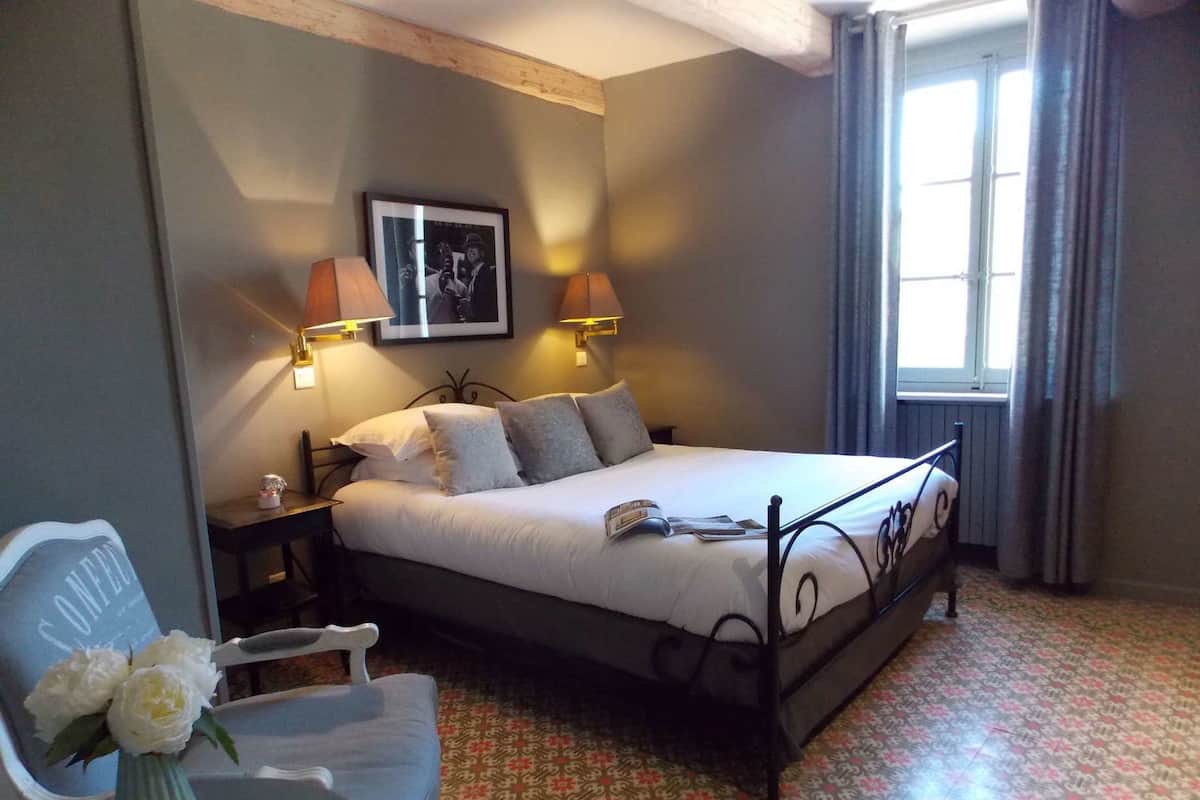 Quarto arrumado com uma cama, duas luminárias de cabeceira, uma fotografia emoldurada na parede, uma cadeira e uma mesa com um vaso de flores.