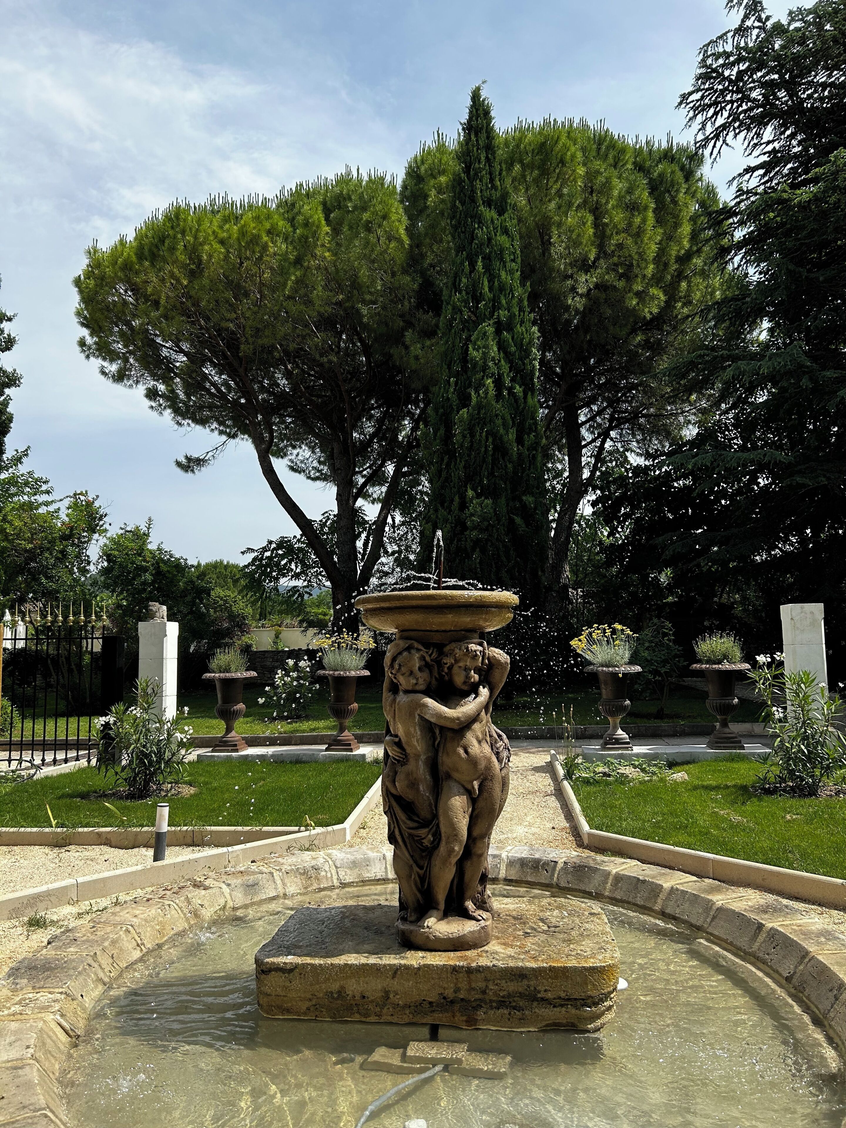 Una fuente con una estatua de dos figuras, rodeada por una balsa de piedra circular y un césped con árboles al fondo.