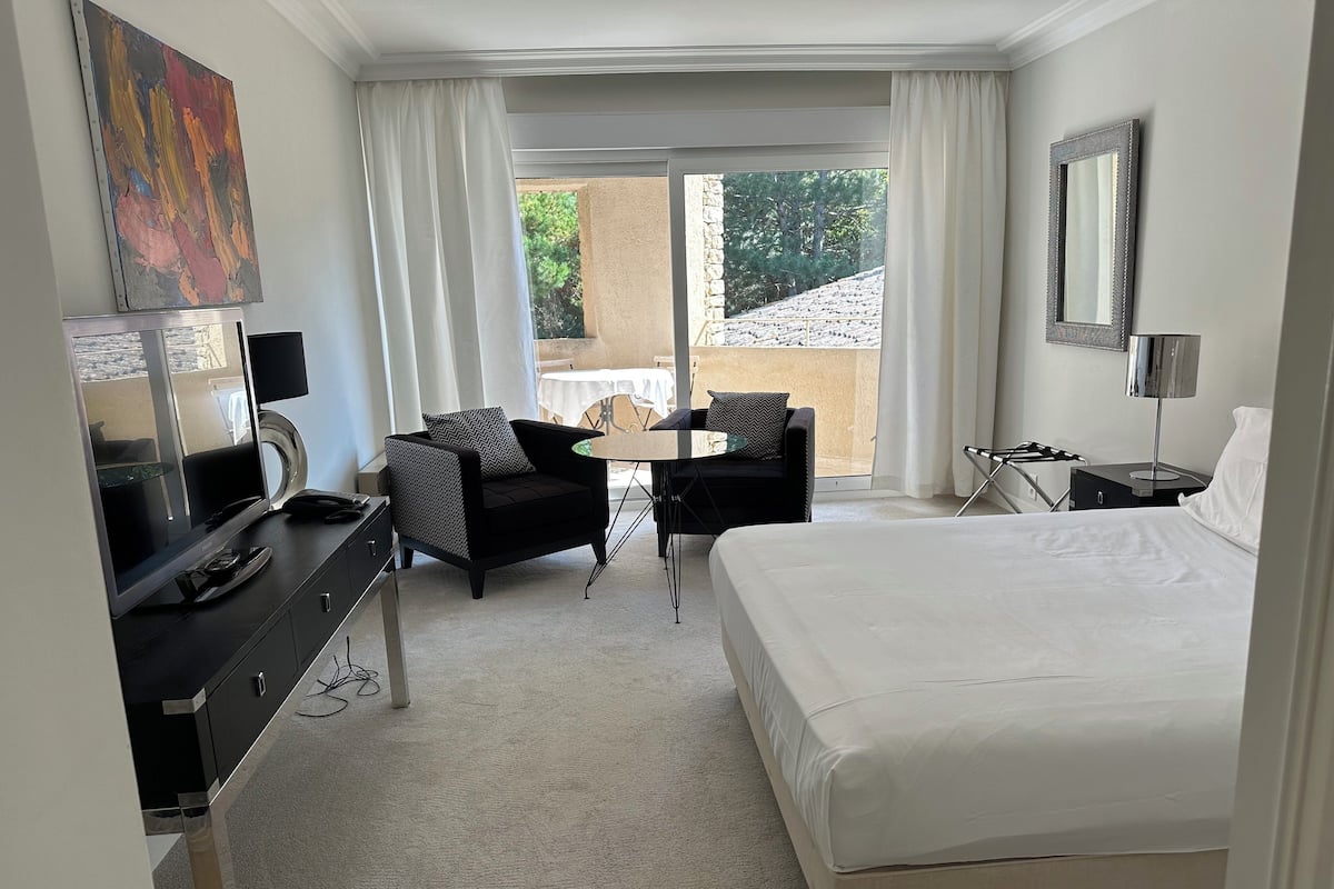 Quarto de hotel com uma cama, uma mesa com televisão, uma cadeira, uma mesinha e uma varanda com vista para árvores.