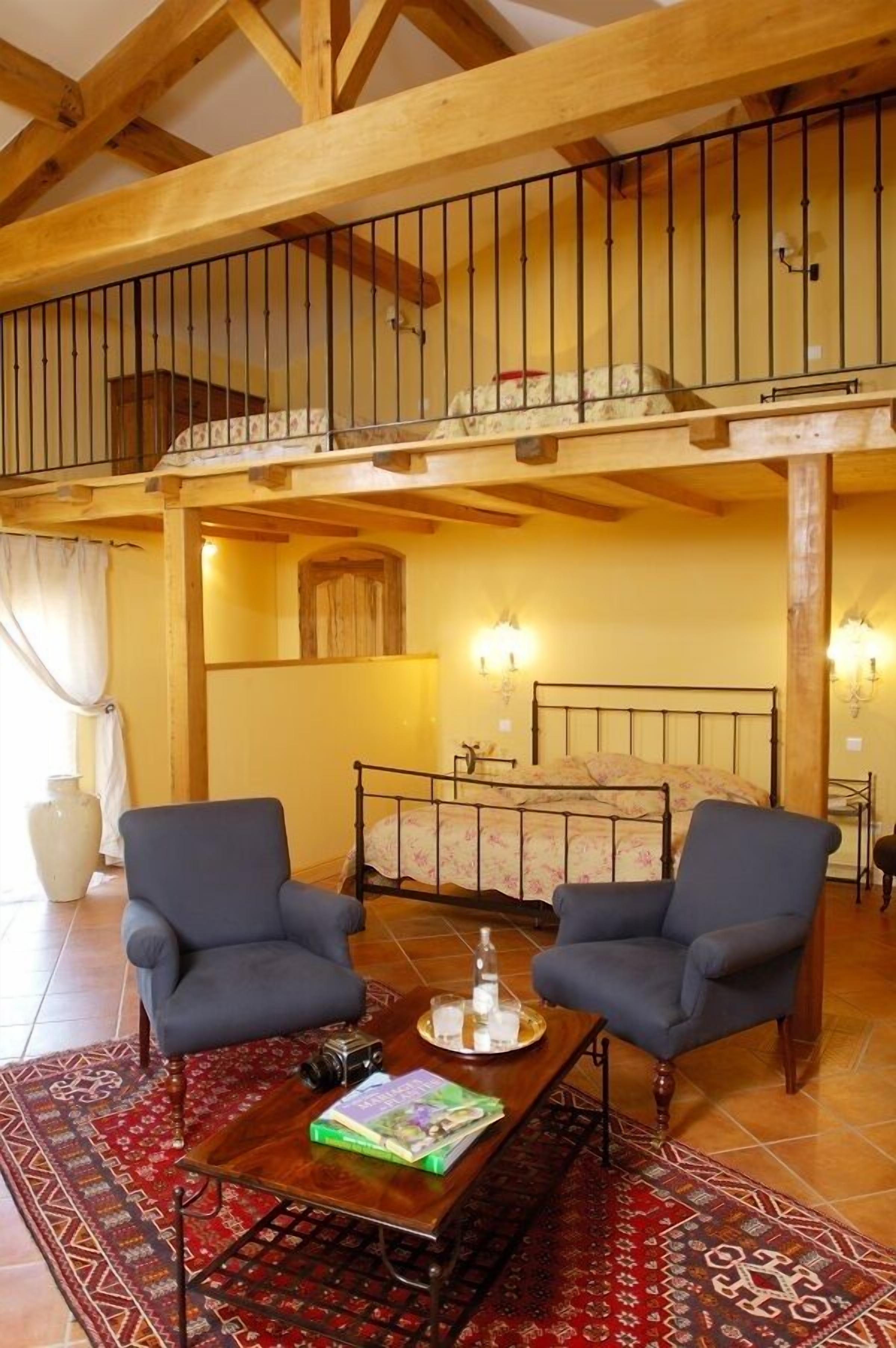 Una habitación con techo de madera, un balcón con barandilla, una cama con colcha estampada, dos sillones, una mesa de centro con libros y una botella, y una alfombra con diseño.
