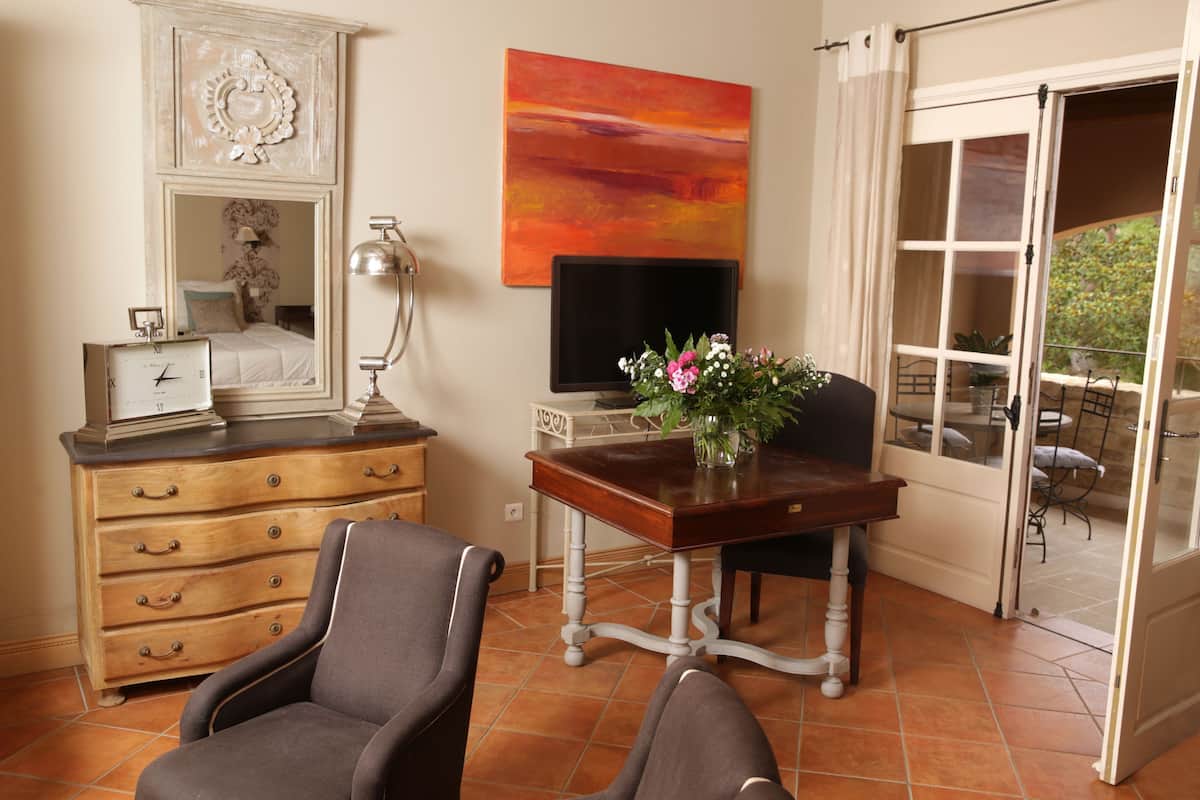 Quarto com cômoda de madeira, escrivaninha com vaso de flores, cadeira, televisão e porta de vidro deslizante que dá para área externa.
