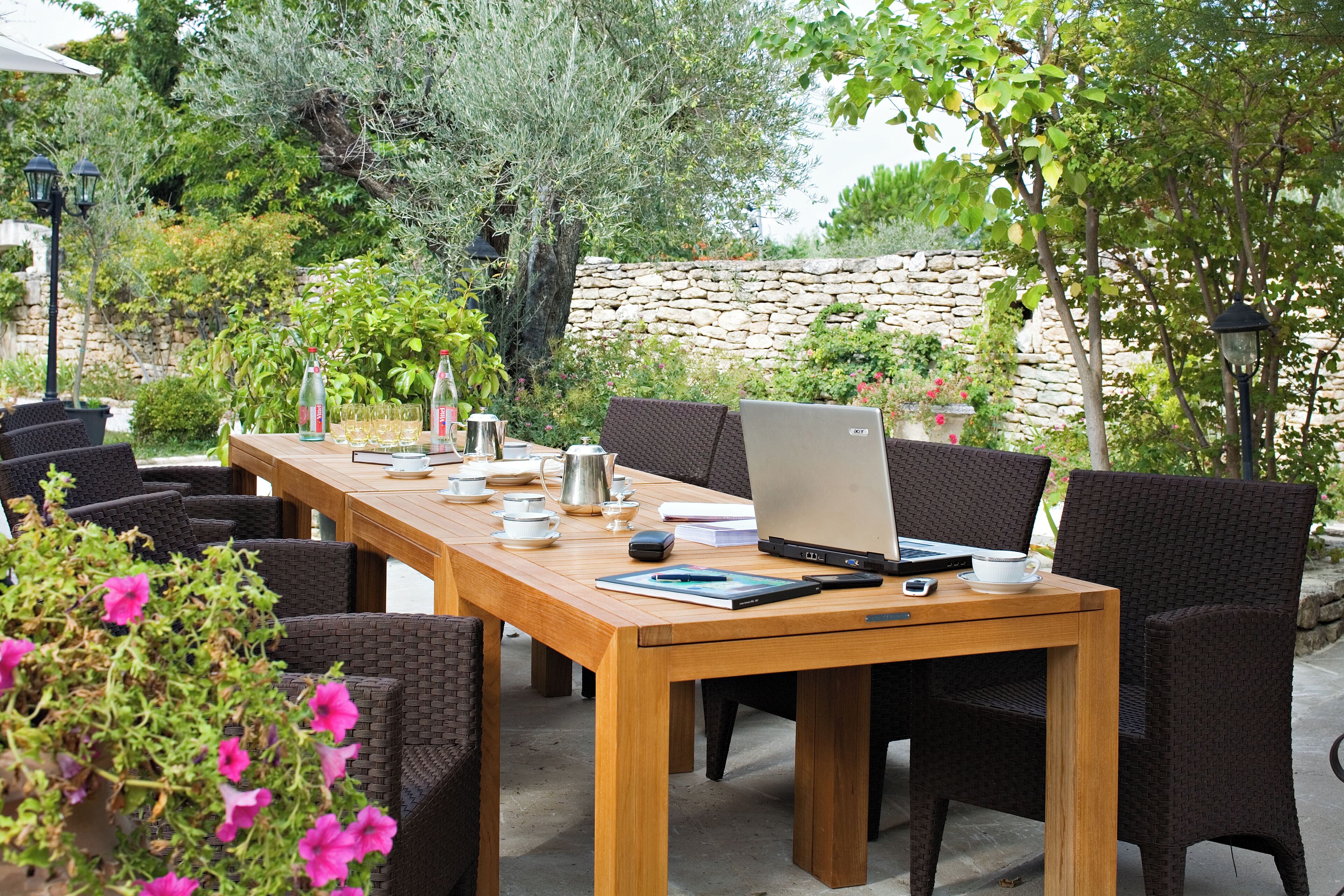 Une table en bois placée à l’extérieur, sur laquelle se trouvent un ordinateur portable, une cafetière, ainsi que différentes tasses et soucoupes.