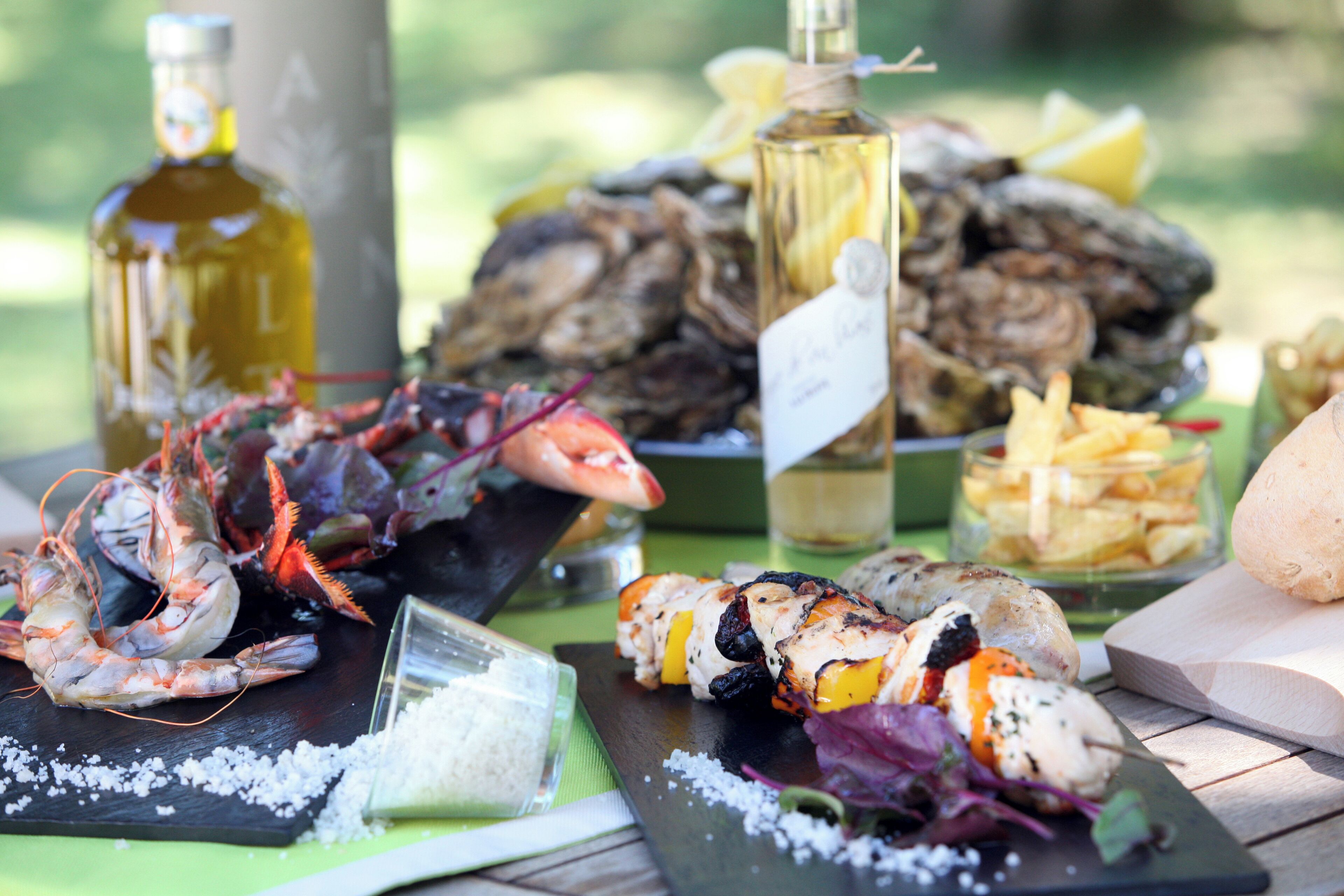 Una mesa con mariscos, incluyendo camarones, ostras y brochetas de pollo a la parrilla, además de botellas de vino y un tazón de papas fritas.