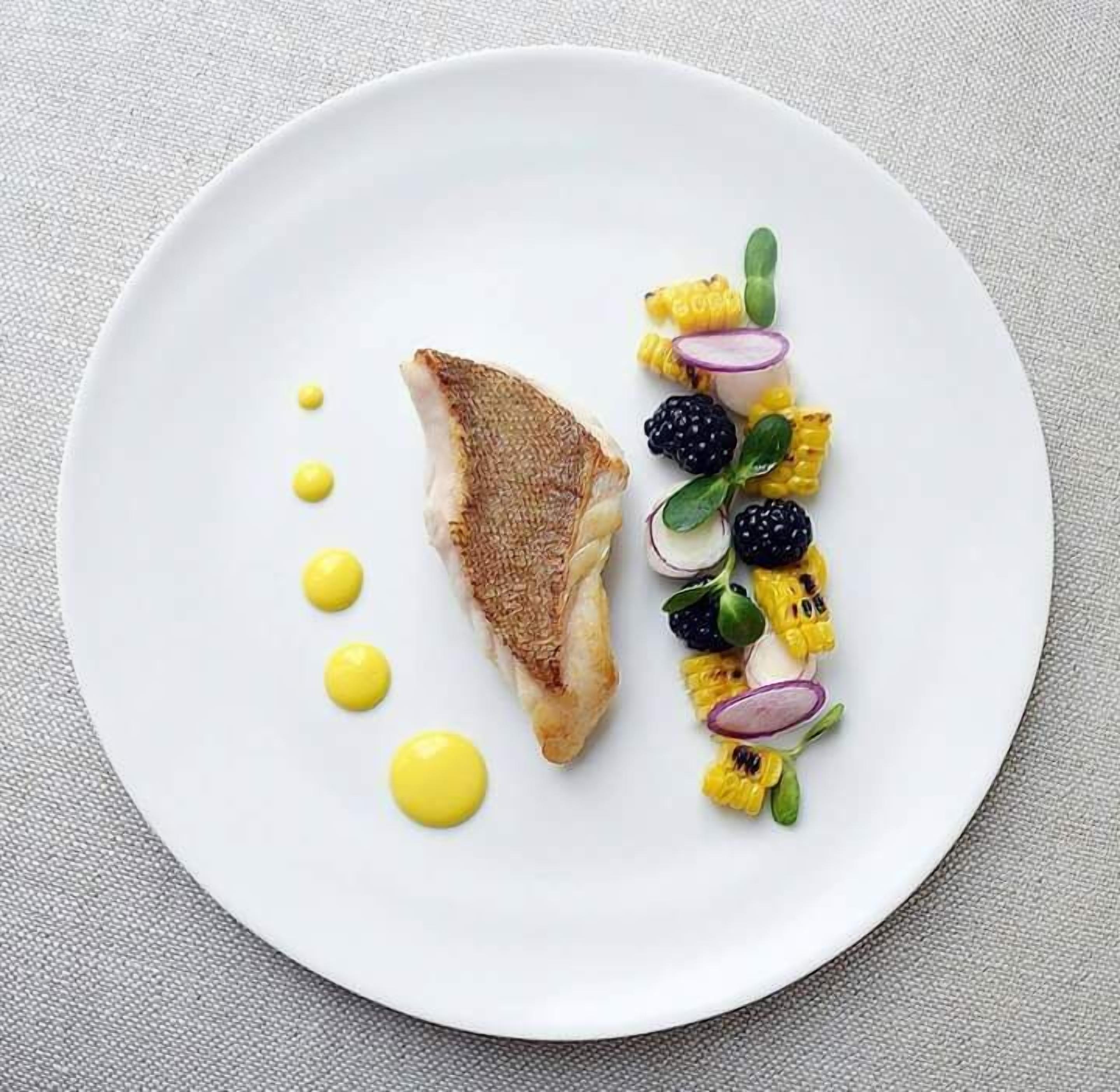 A white plate with a piece of grilled fish, grilled corn, blackberries, and a yellow sauce.