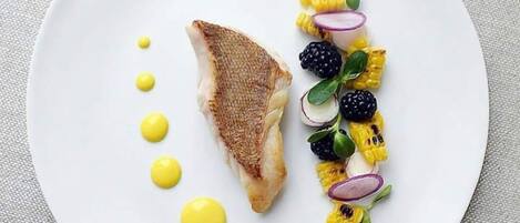 A white plate with a piece of grilled fish, grilled corn, blackberries, and a yellow sauce.