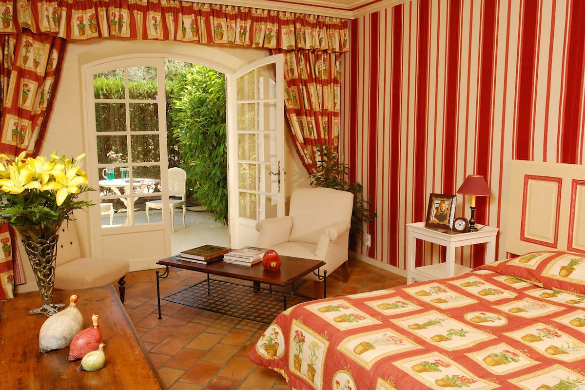 Quarto com cama, mesa de centro, cadeira, mesa lateral e vista para um jardim por uma porta aberta.