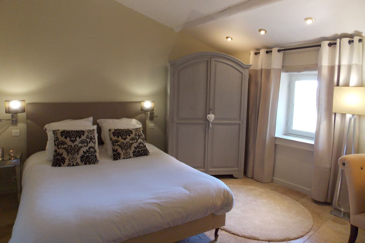 Quarto com uma cama, duas luminárias de cabeceira, um guarda-roupa, uma janela com cortinas e uma cadeira.