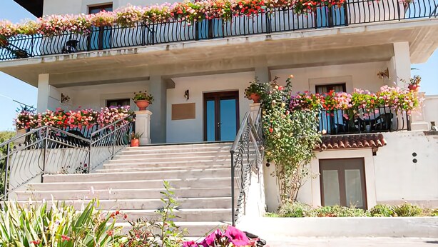 A two-story building with a balcony adorned with flowers, a staircase leading to the entrance, and a flower pot with purple flowers in the foreground. - Albergo della Corte (Pescasseroli)