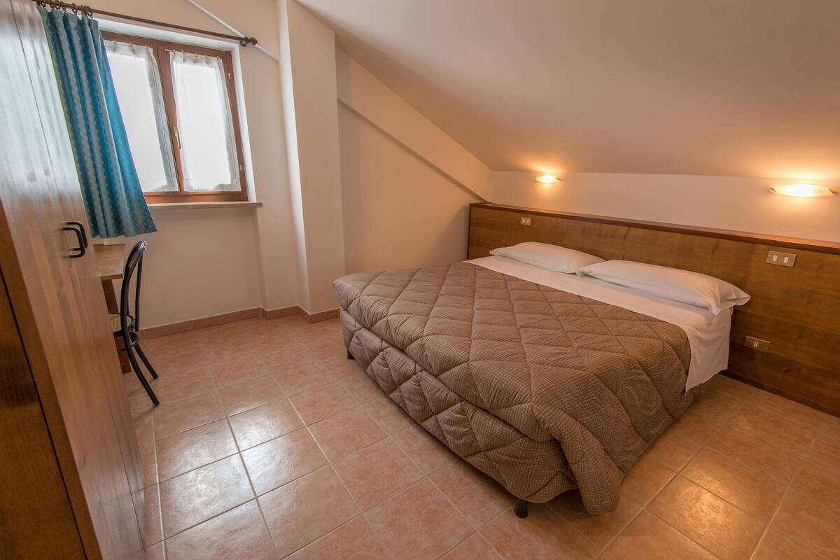 A bedroom with a bed, a window with curtains, a headboard with lights, and a tiled floor.