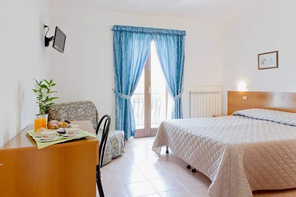 A hotel room with a bed, a desk with breakfast, a chair, and a window with curtains.