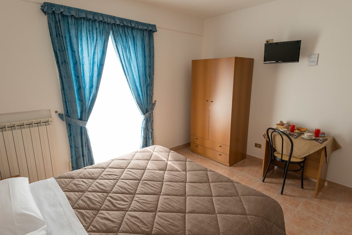 A bedroom with a bed, a wardrobe, a desk with a chair, and a television on the wall.