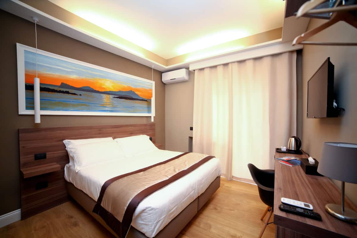 A hotel room with a bed, a desk with a lamp, a TV, a painting of a sunset, and an air conditioning unit.
