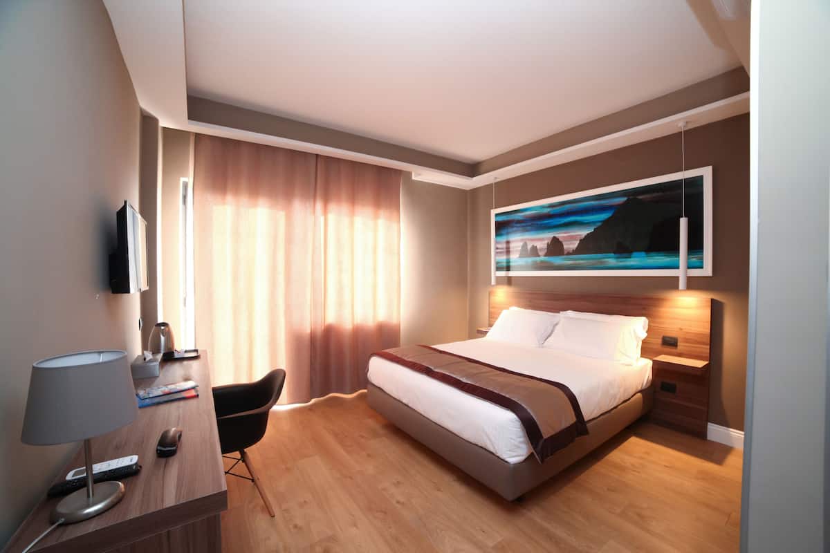 A modern hotel room with a large bed, a desk with a lamp, a chair, and a painting on the wall.