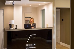 Un comptoir d’accueil avec un ordinateur portable, un téléphone et un vase de fleurs. On peut lire l’inscription « CHAMBRES PRIVILÉGIÉES, 7e ÉTAGE ».