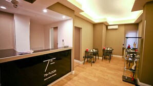 A hotel reception area with a black counter labeled "7th FLOOR EXCLUSIVE ROOMS," a dining area with a table and chairs, and a display shelf with decorative items. - 7th Floor Suite (Naples)