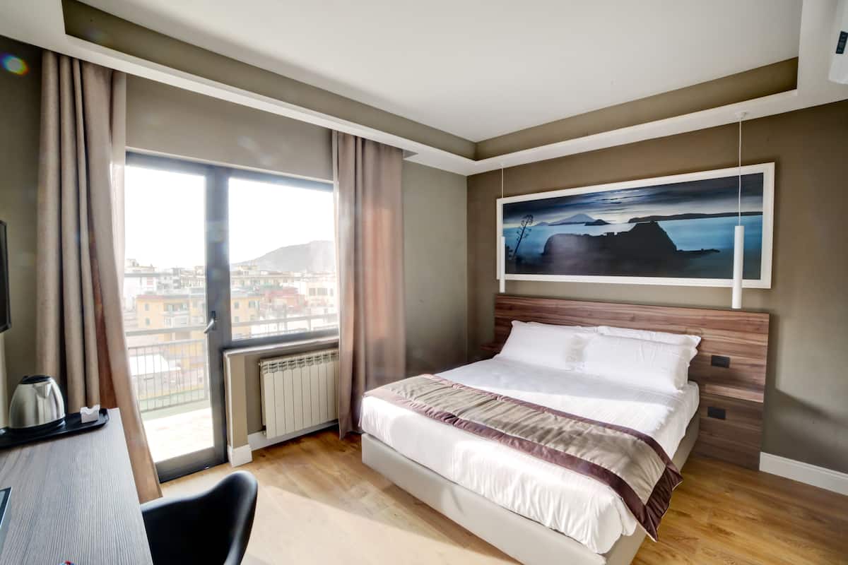 A hotel room with a large bed, a desk with a kettle, a view of buildings through a window, and a painting on the wall.