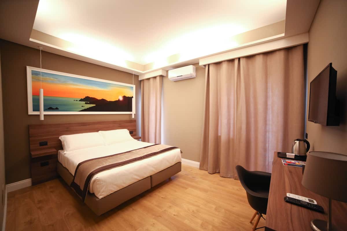 A hotel room with a bed, a desk, a chair, a television, and a painting on the wall.