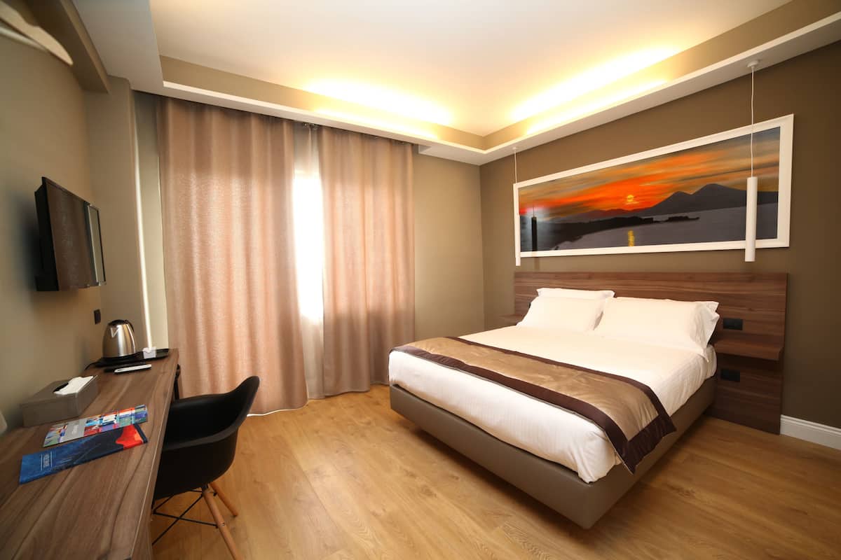 A hotel room with a bed, a desk with a chair, a TV, and a painting on the wall.