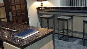 Una cocina con encimera de granito, estufa y una pequeña isla con un libro encima. Hay dos taburetes de bar y una ventana con persianas.