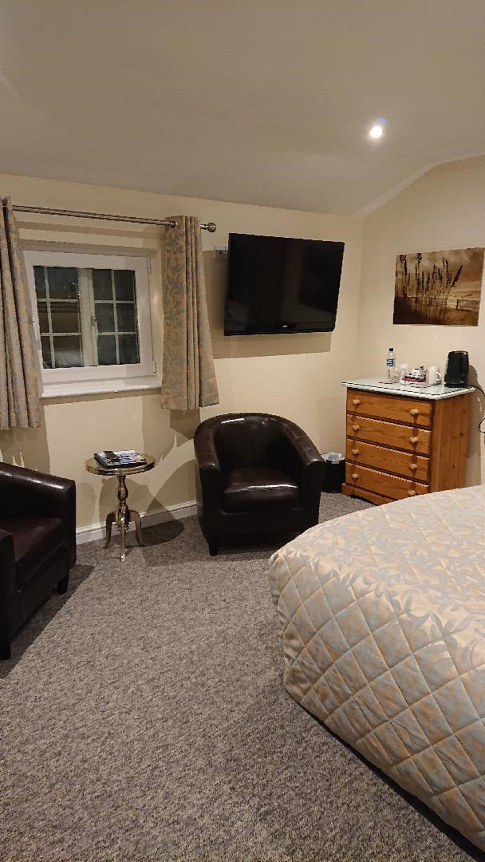 A hotel room with a bed, a chair, a small table, a dresser, a television, and a window with curtains.