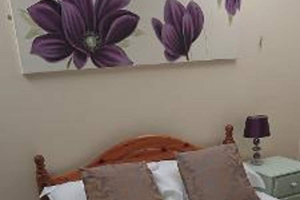 A bedroom with a wooden bed, white bedding, two pillows, a nightstand with a lamp, and a wall-mounted painting of purple flowers.