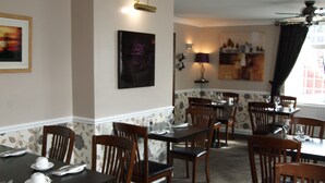 Un comedor con mesas y sillas de madera, obras de arte en las paredes y un ventanal con cortinas.