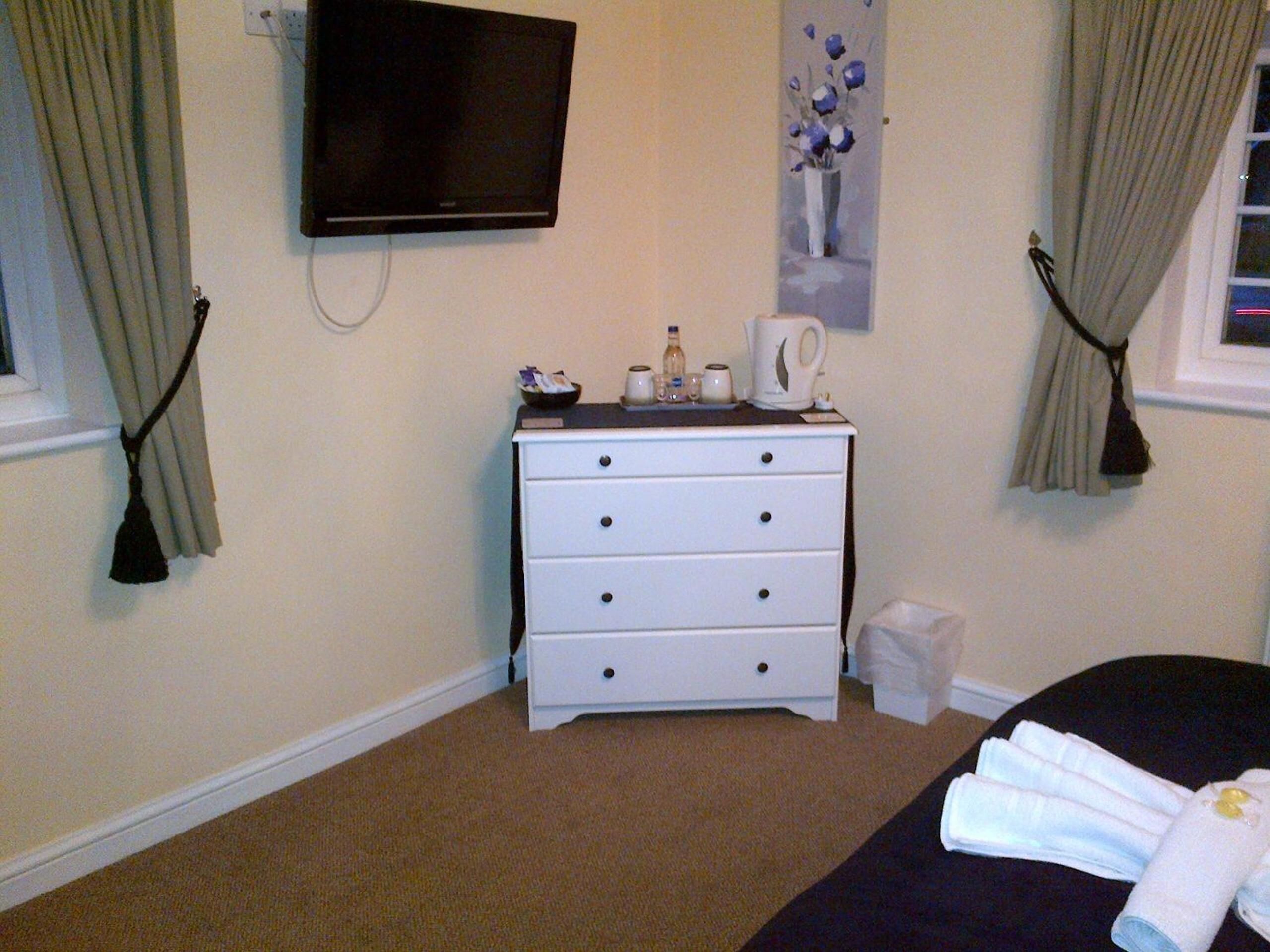 A hotel room with a bed, a white dresser with drawers, a flat-screen TV mounted on the wall, a vase with flowers, and a window with curtains.