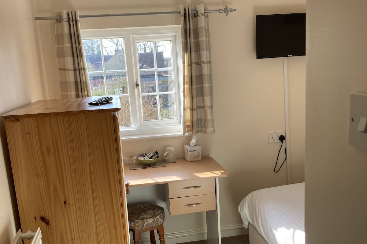 A bedroom with a wooden wardrobe, a desk with a chair, a bed, a radiator, a TV, and a window with curtains.
