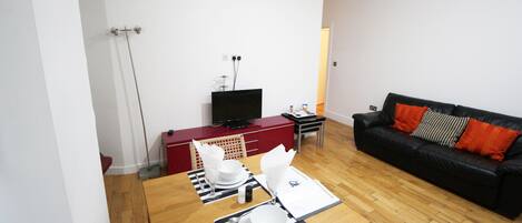 Un comedor con una mesa de madera dispuesta para dos personas, un sofá de cuero negro con cojines naranjas, y un televisor sobre un mueble rojo.