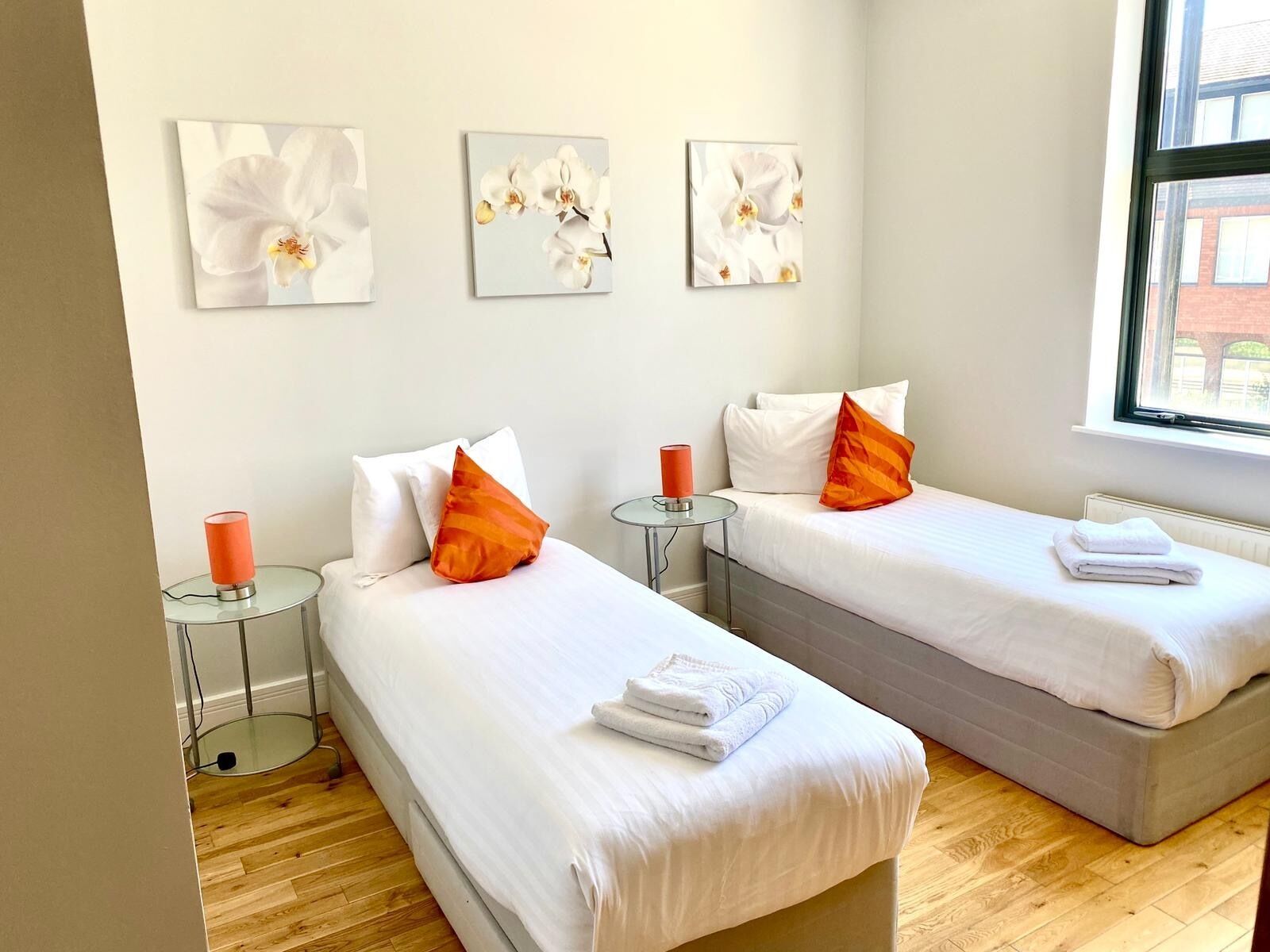 A hotel room with two beds, white bedding, orange pillows, a glass table with a red candle, and orchid paintings on the wall.