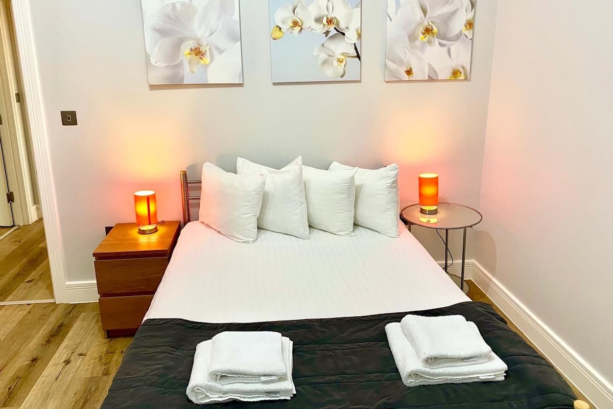 Een hotelkamer met een bed, kussens, een nachtkastje met een lamp, een klein rond tafeltje met een lamp, en drie orchideeënschilderijen aan de muur.