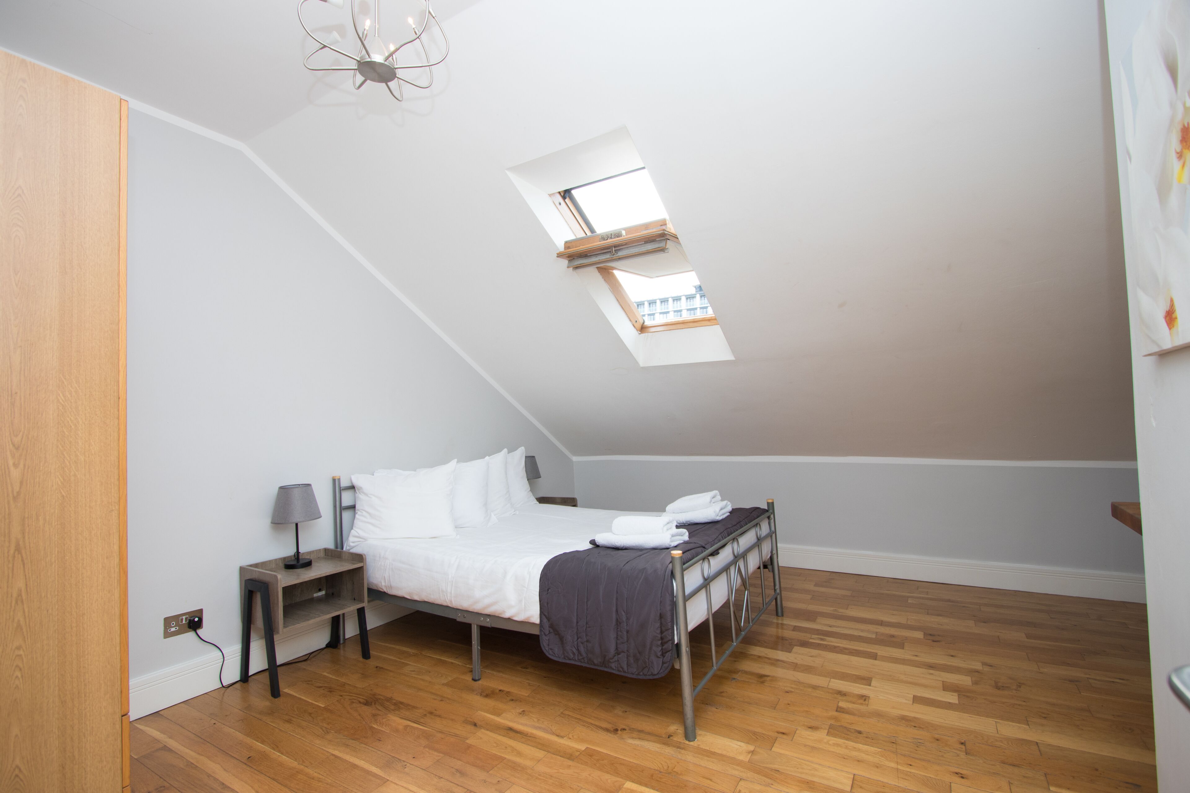 A bedroom with a sloped ceiling, a skylight, a bed, a nightstand, and a wooden floor.