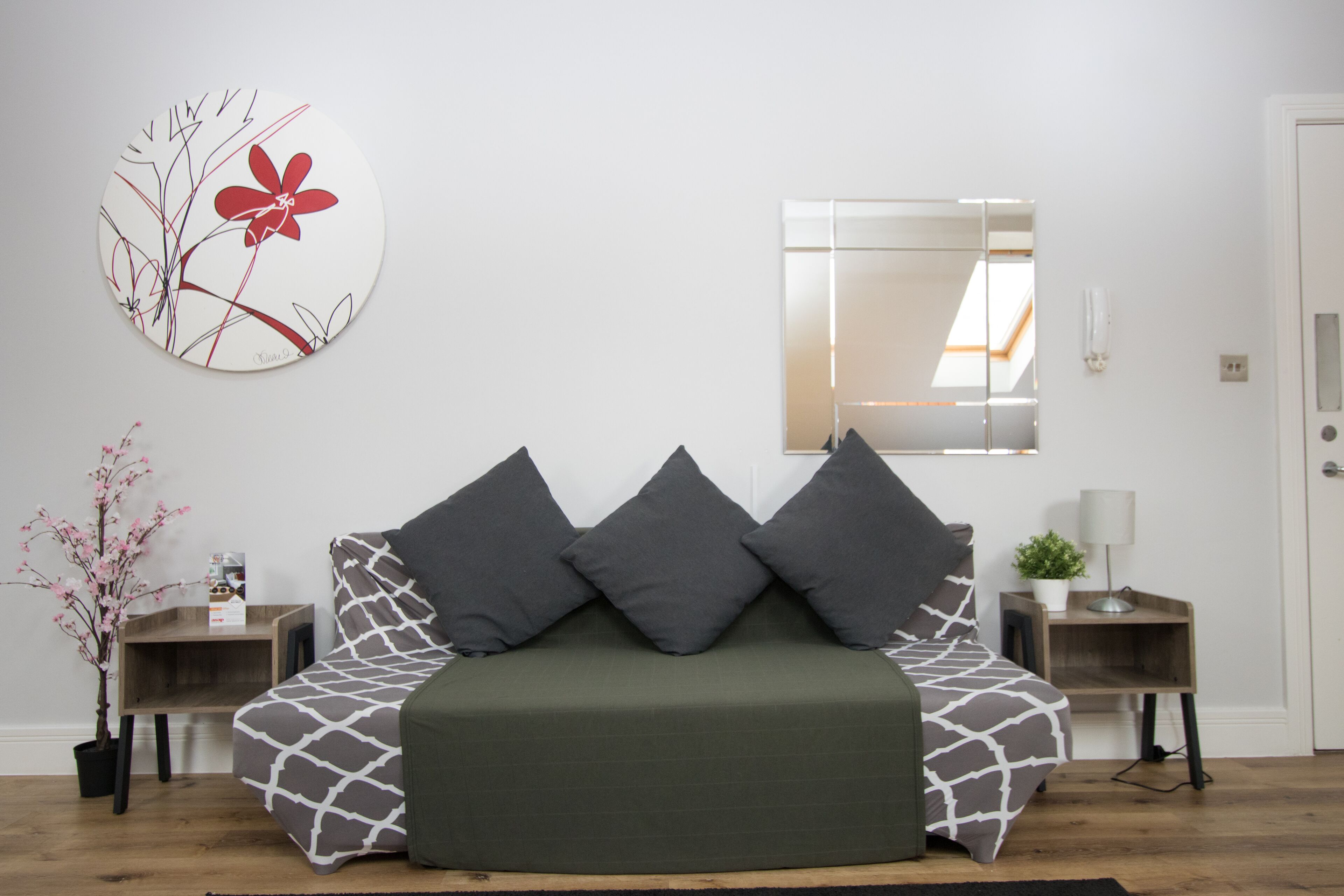 Une chambre moderne avec un canapé-lit vert, des coussins à motifs gris et blancs, une œuvre d’art murale ronde, un miroir et une petite plante posée sur une table de chevet en bois.