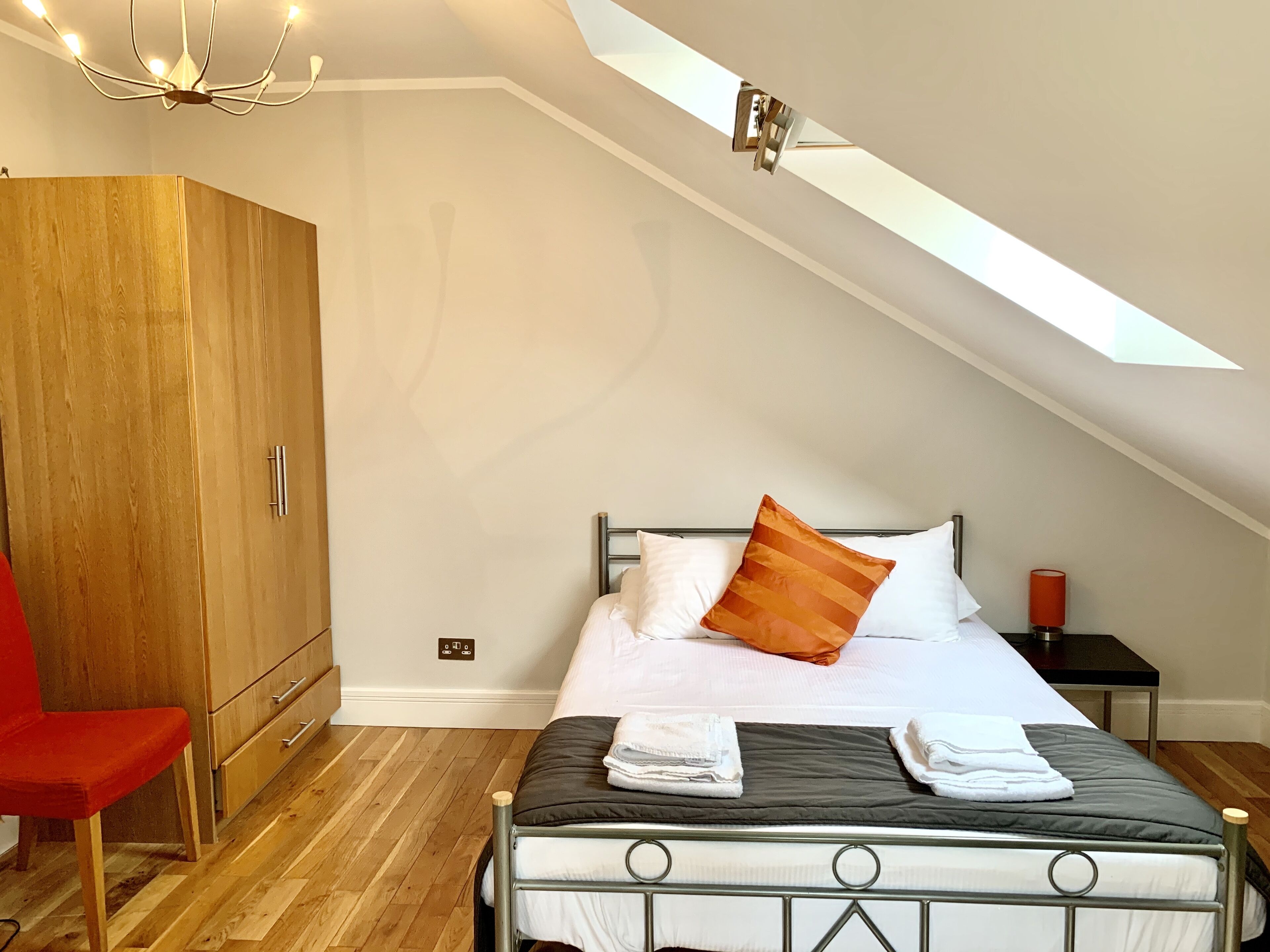 A bedroom with a sloped ceiling, a bed with pillows, a wooden wardrobe, a red chair, and a nightstand with a lamp.