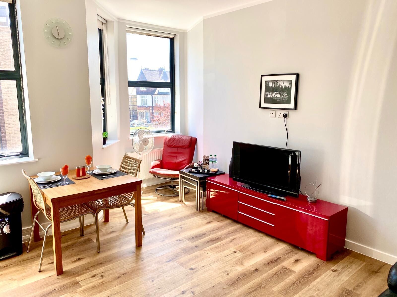 A modern living room with a wooden dining table, a red chair, a TV on a red stand, and a window with a view of buildings.