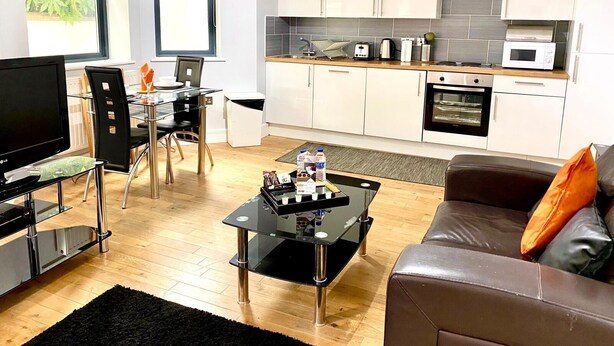 Eine moderne Küche mit weißen Schränken, einer Mikrowelle und einem Backofen. Es gibt einen Essbereich mit einem Glastisch und Stühlen, sowie ein Wohnzimmer mit einem Ledersofa und einem Couchtisch.