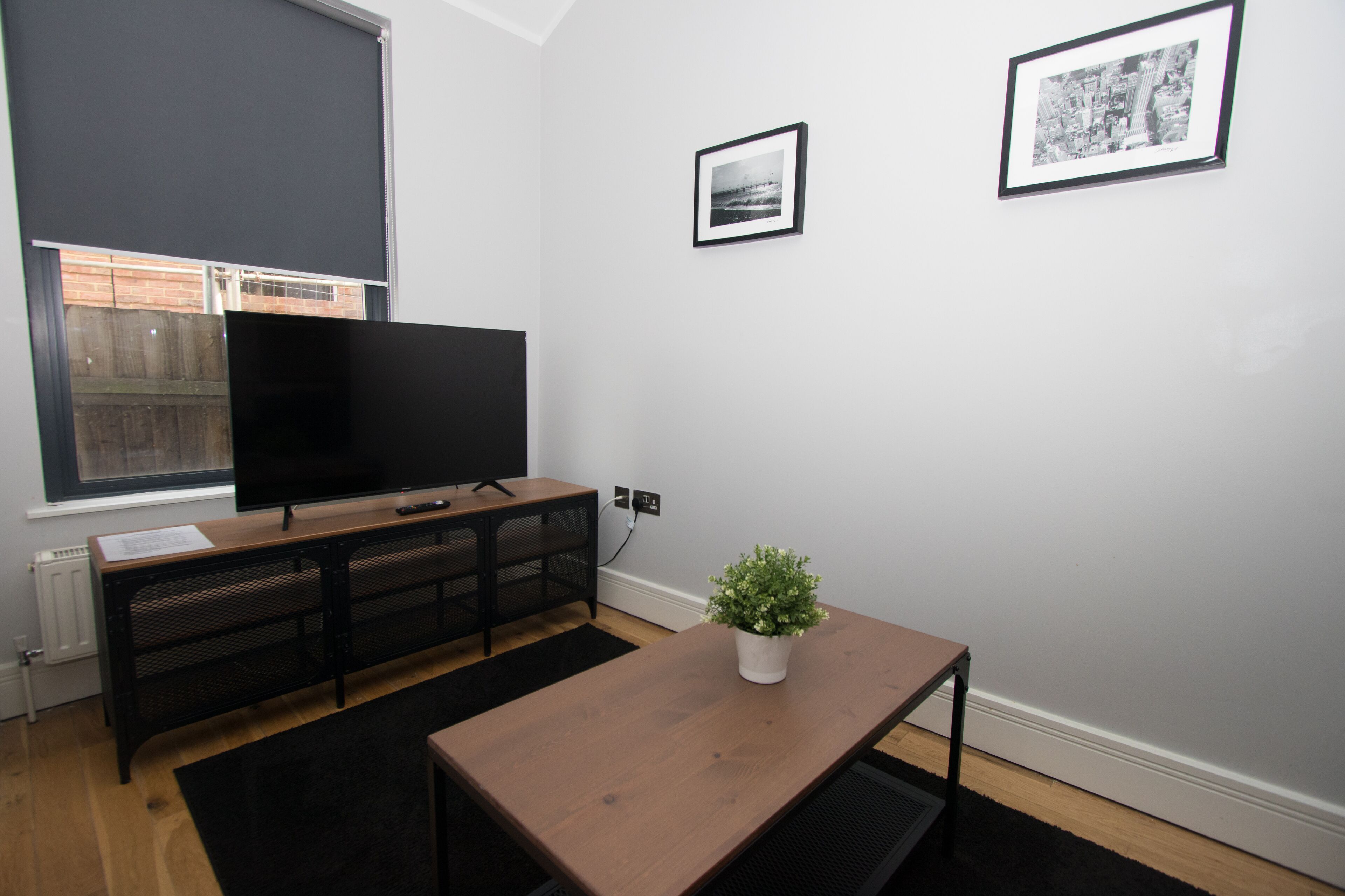 A modern living room with a flat-screen TV, a wooden coffee table, a potted plant, and two framed pictures on the wall.