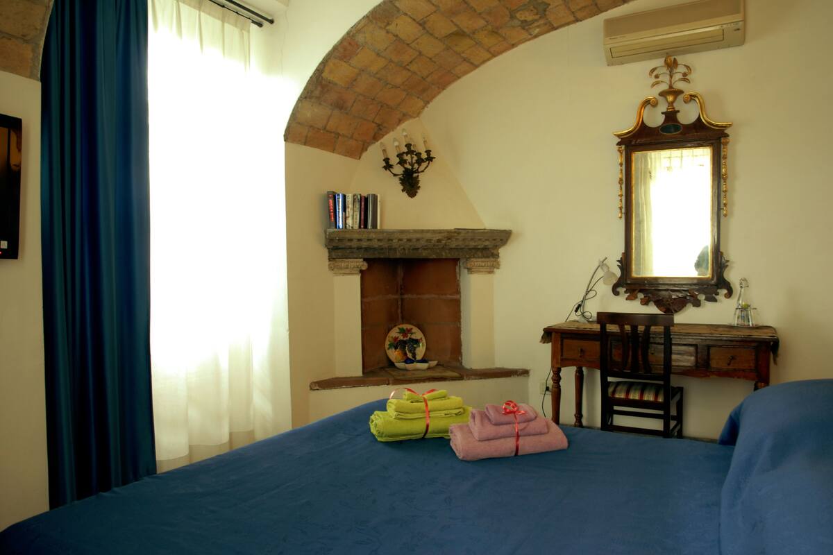A bedroom with a bed, a desk, a chair, a fireplace, and a mirror.