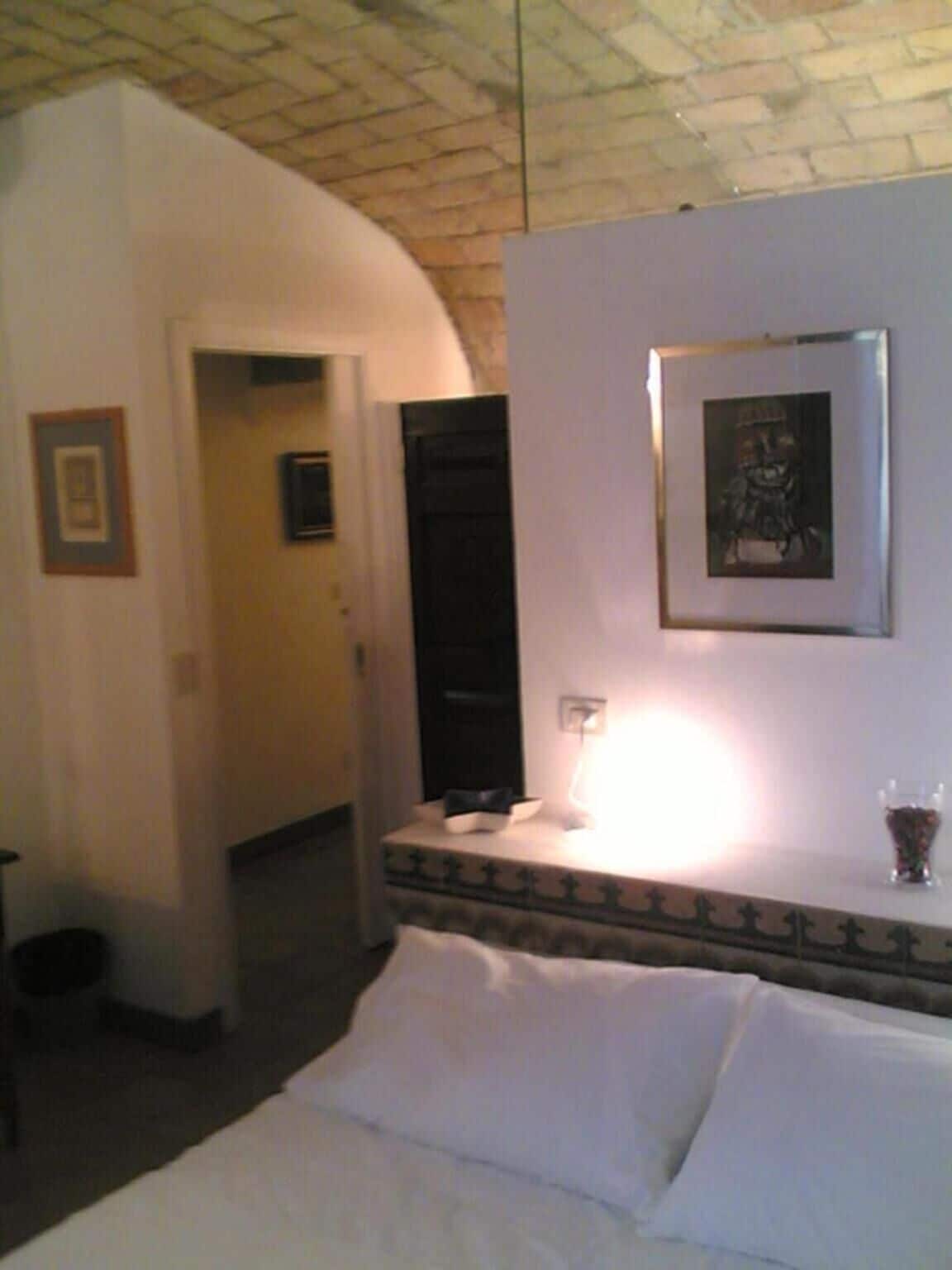 Una habitación de hotel con una cama, una mesita de noche con una lámpara, un cuadro enmarcado en la pared y una puerta que da a otra habitación.