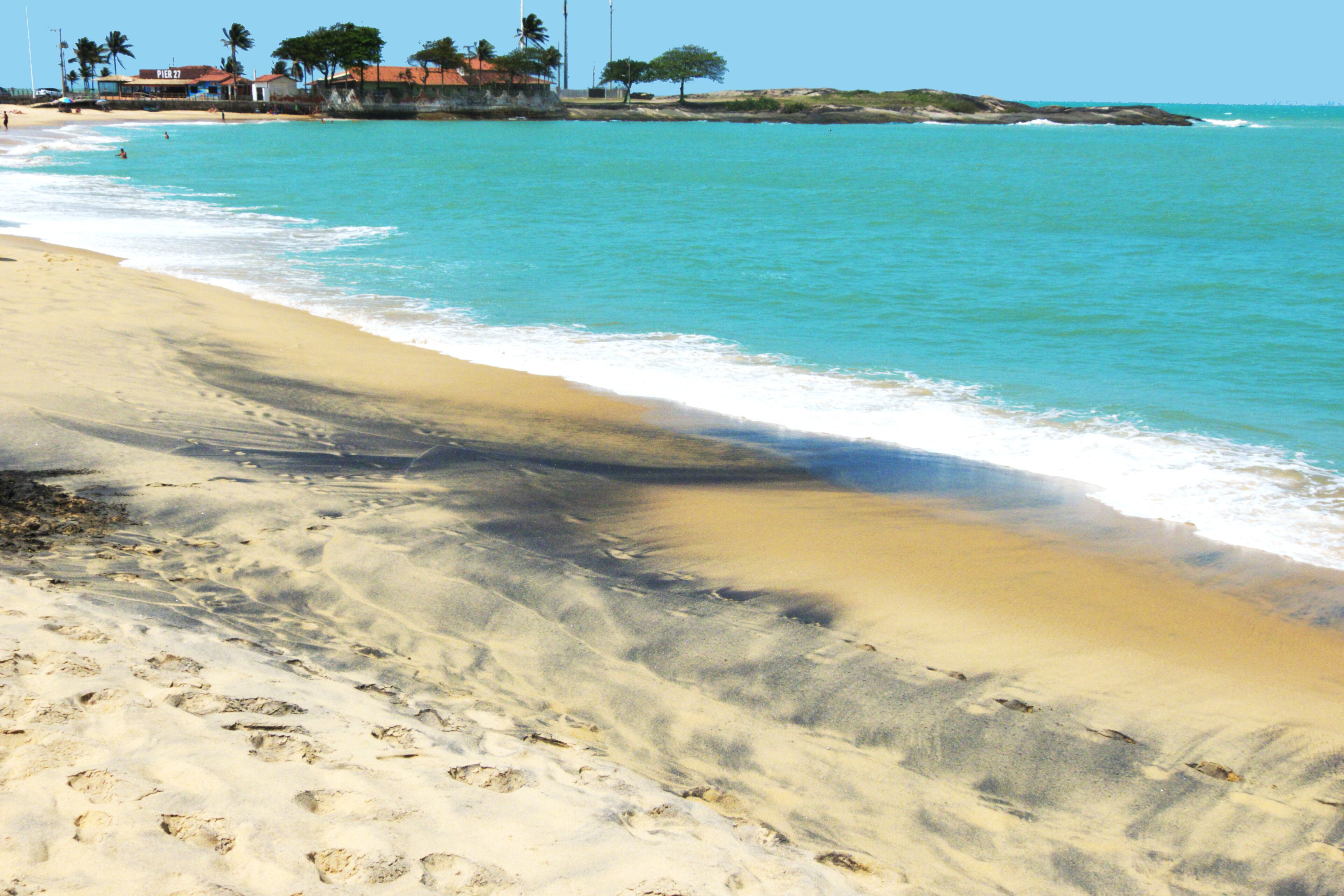 A sandy beach with footprints leading to the water, a clear sky, and buildings in the background.
