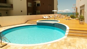 Une piscine sur le toit, dotée de chaises longues et de parasols, entourée d’une terrasse en bois et d’un bâtiment avec des balcons.