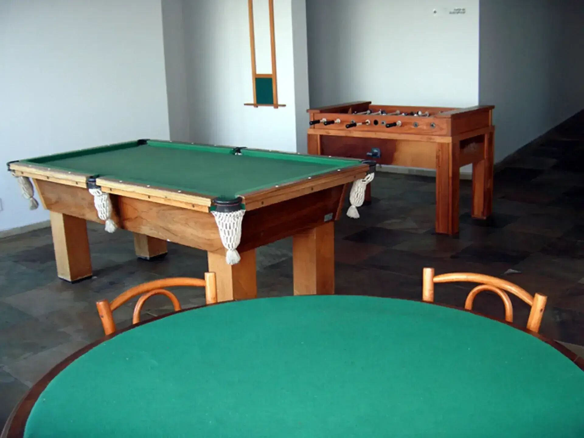 A pool table with a green felt surface, a foosball table, and a wooden chair.