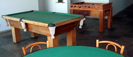A pool table with a green felt surface, a foosball table, and a wooden chair.