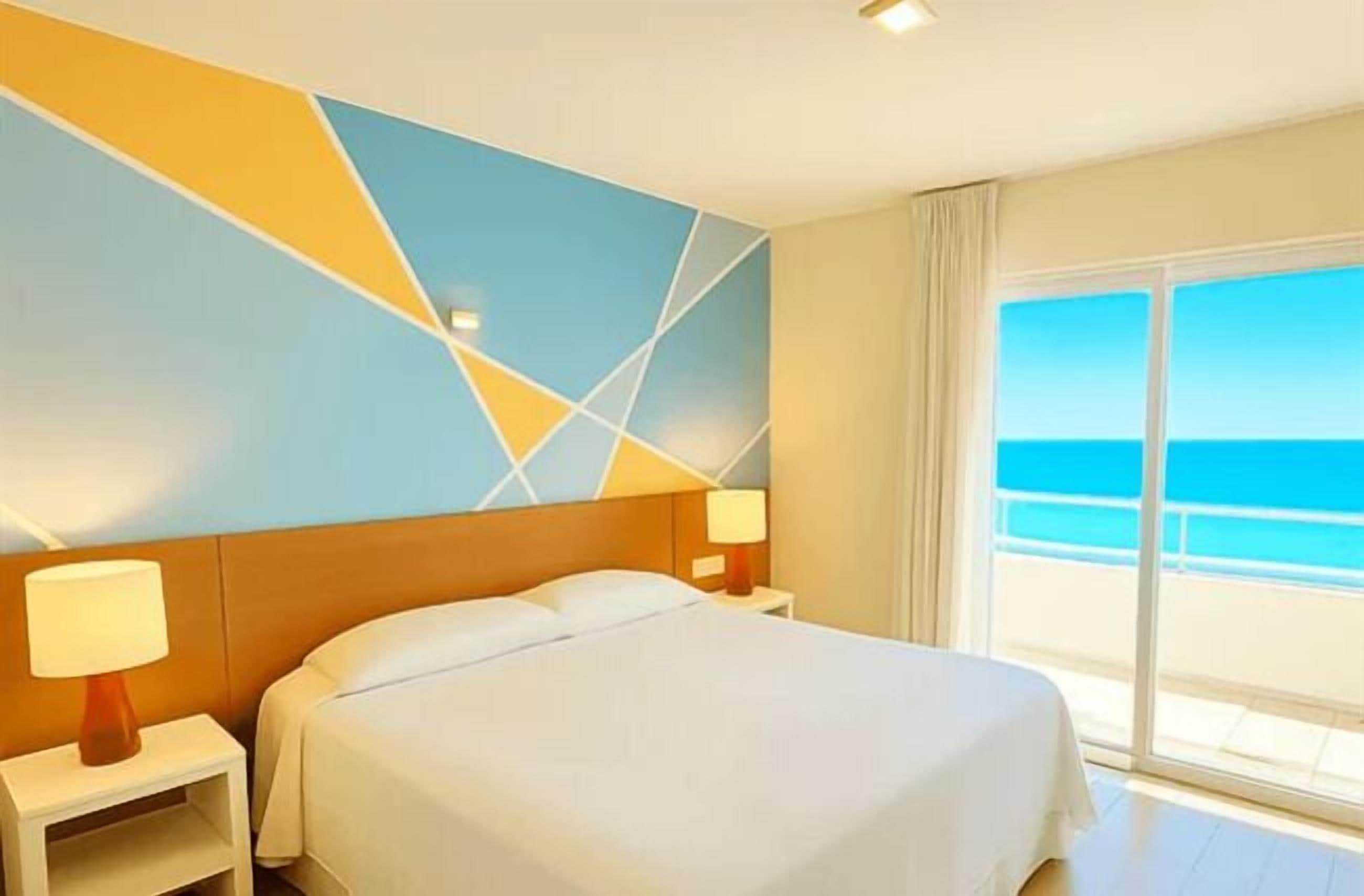 A hotel room with a bed, two bedside tables with lamps, a large window with a view of the ocean, and a geometric wall design.