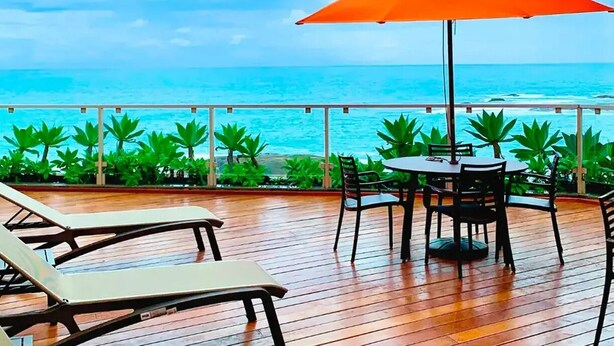 Une terrasse en bois avec une table et des chaises, un parasol orange et une vue sur l’océan.